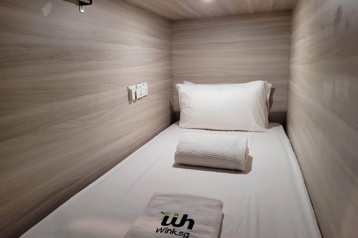 Quarto de hotel pequeno e moderno, com uma cama de solteiro, mesa de cabeceira e luminária fixada na parede.