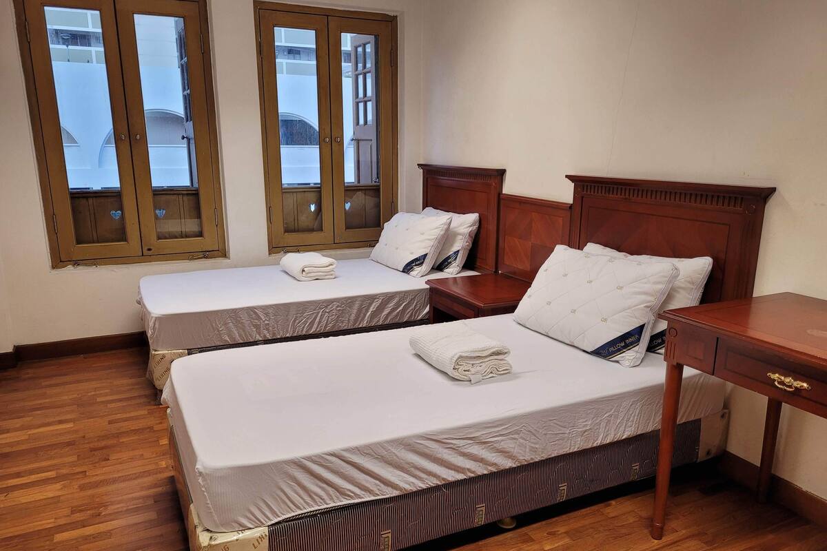 Quarto de hotel com duas camas de solteiro, cabeceiras de madeira, uma mesinha de madeira e uma janela com vista para edifícios.