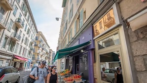 A street with a fruit stand, buildings, and parked cars. - Luxurious apartment, wifi, air conditioning, 50M from the beach (Nice)