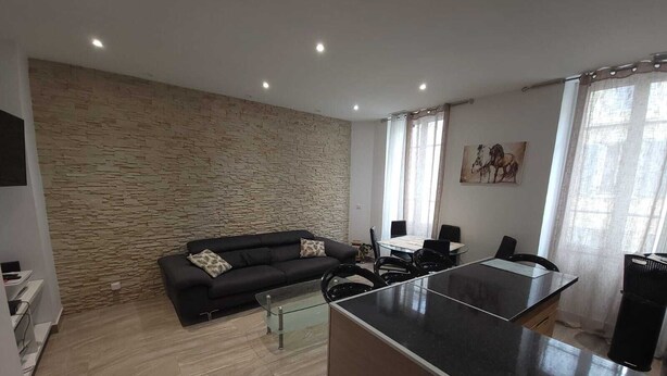A modern living room with a stone wall, a black sofa, a glass coffee table, and a dining area with a table and chairs. - Luxurious apartment, wifi, air conditioning, 50M from the beach (Nice)