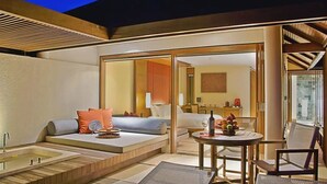 A modern hotel room with a large sliding glass door leading to a balcony, a sofa with cushions, a small round table with a bottle and fruit, and a bathtub.