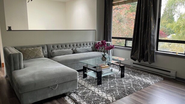 A modern living room with a grey sectional sofa, a glass coffee table, and a view of trees through a window. - Remodel Spacious Modern Retreat (Shoreline)