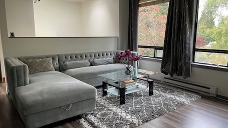 A modern living room with a grey sectional sofa, a glass coffee table, and a view of trees through a window.