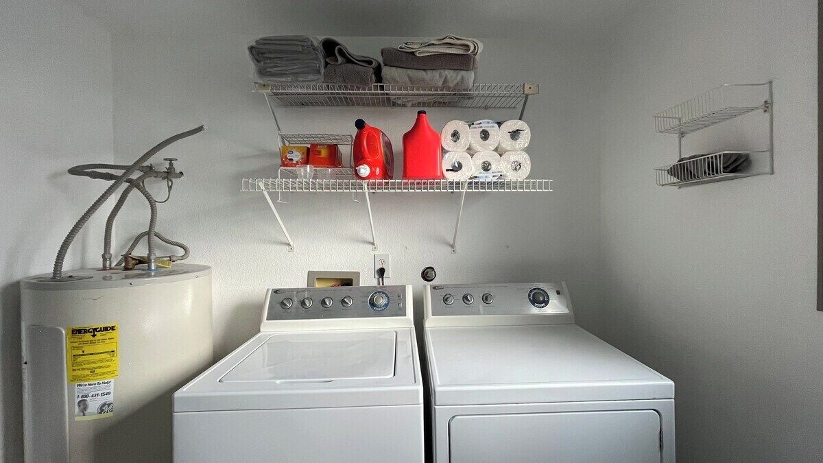 Un área de lavandería con dos lavadoras blancas, un calentador de agua y estantes con toallas y productos de limpieza.