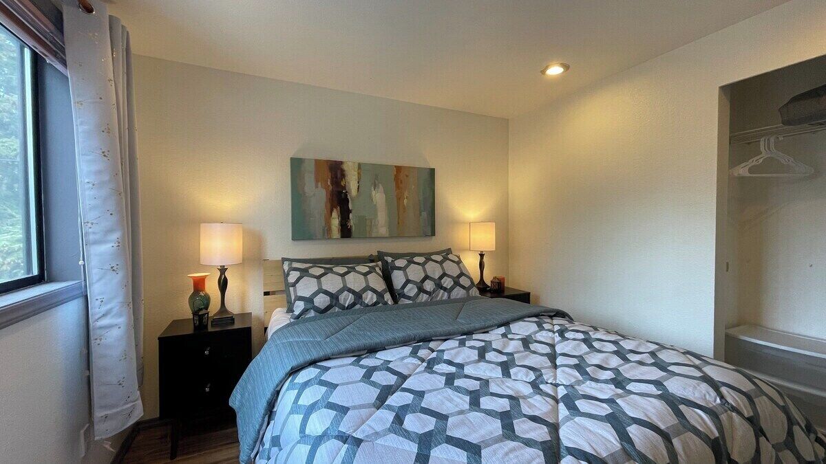 A bedroom with a bed, two bedside tables, a wall-mounted artwork, and a window with curtains.