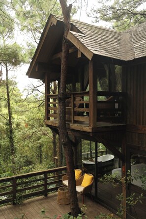 A wooden treehouse with a balcony, built into a tree, featuring a small table and chair, surrounded by a wooden deck. - Amaya Retreat (Hanoi)