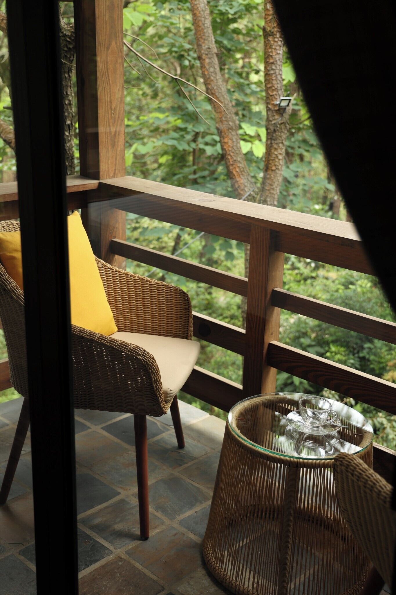 Ein Korbstuhl mit Kissen, ein Glastisch mit einer Tasse und ein Korb auf einem Balkon mit Blick auf Bäume.