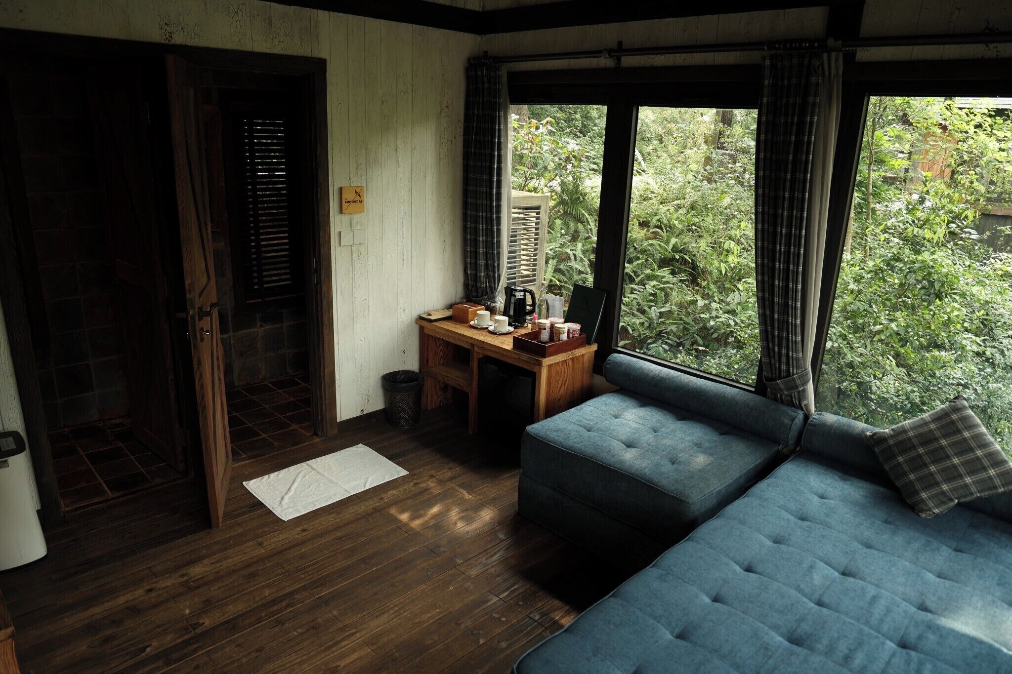 A cozy room with a wooden floor, a blue sofa, a small wooden table with a coffee maker, and a window with curtains.