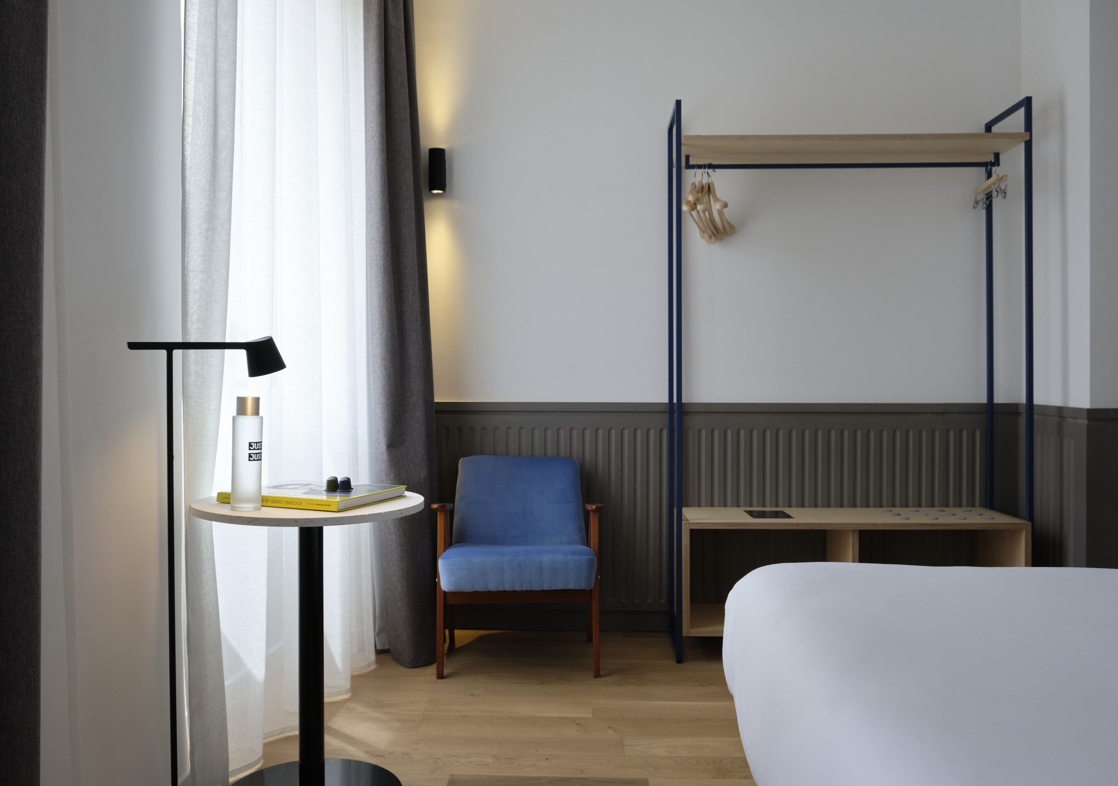A hotel room with a bed, a chair, a small table with a lamp, a bedside table, and a wall-mounted shelf with decorative items.