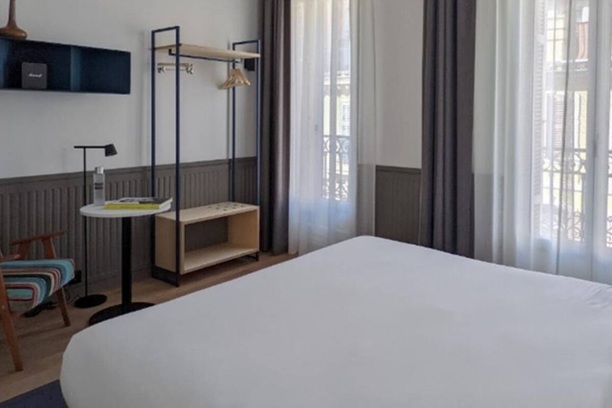 A hotel room with a large bed, a chair, a small table, a shelf with books and a vase, and a window with curtains.