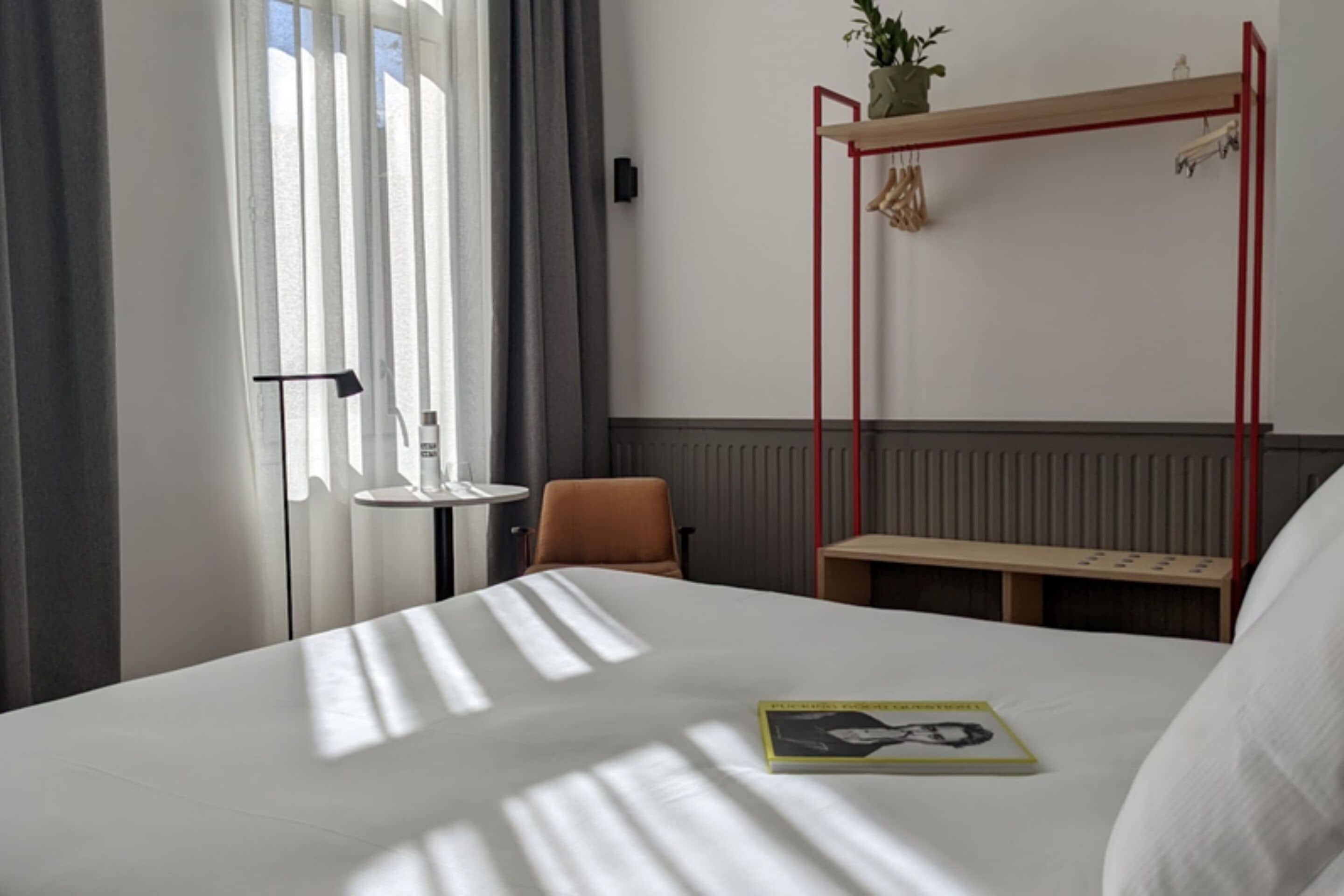 A hotel room with a large bed, a small table, a chair, a red metal shelf with a plant, and a window with curtains.