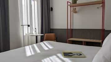 A hotel room with a large bed, a small table, a chair, a red metal shelf with a plant, and a window with curtains.