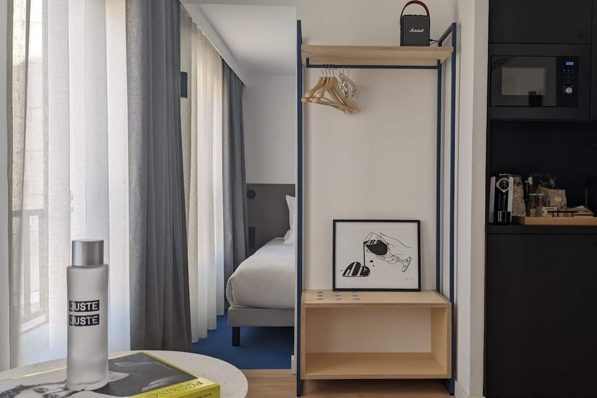 A modern hotel room with a bed, a bedside table with a lamp, a shelf with a framed picture, and a wardrobe with hanging clothes.
