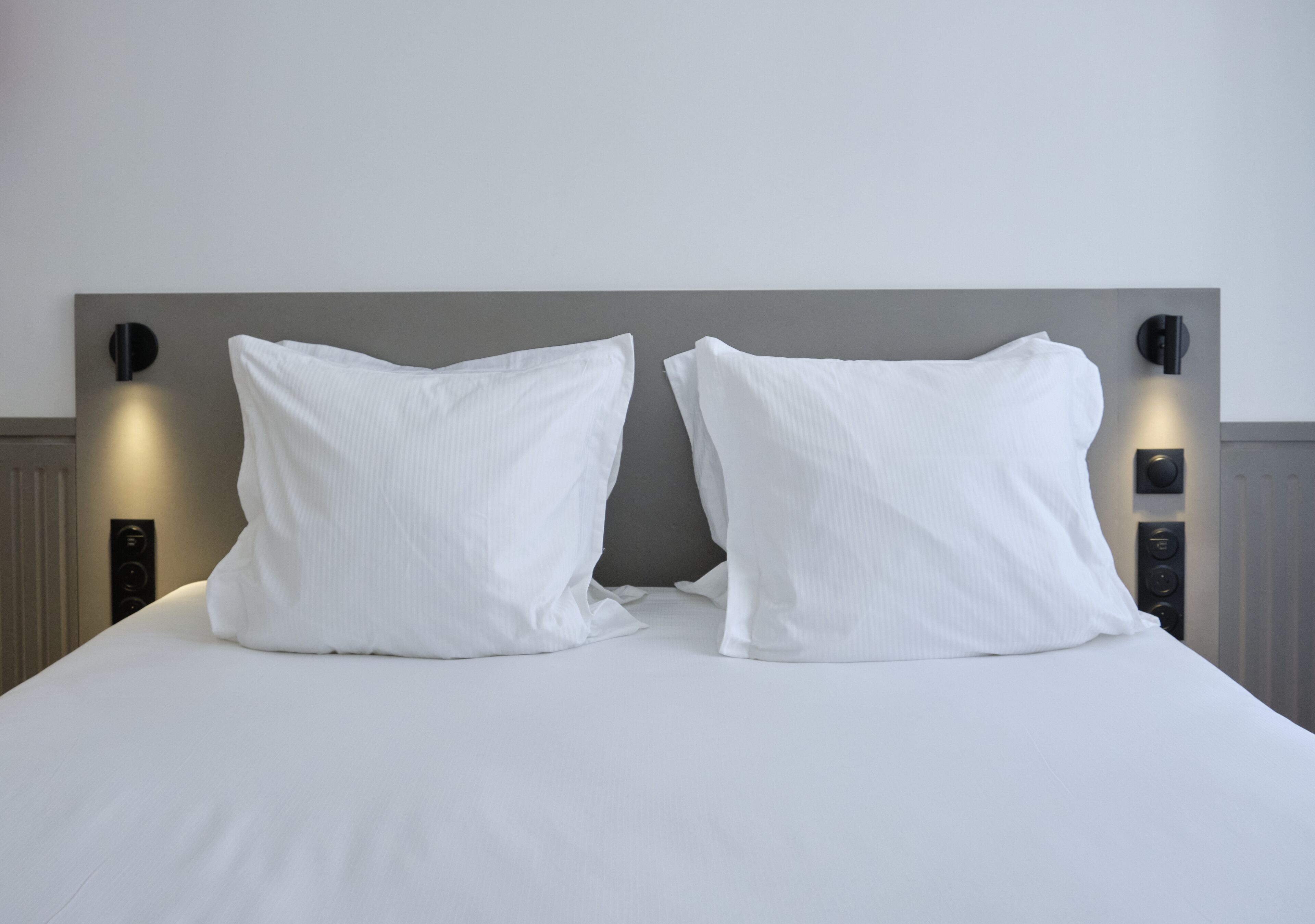 Cama com lençóis e duas almofadas brancas, com luminárias fixadas na parede de cada lado.