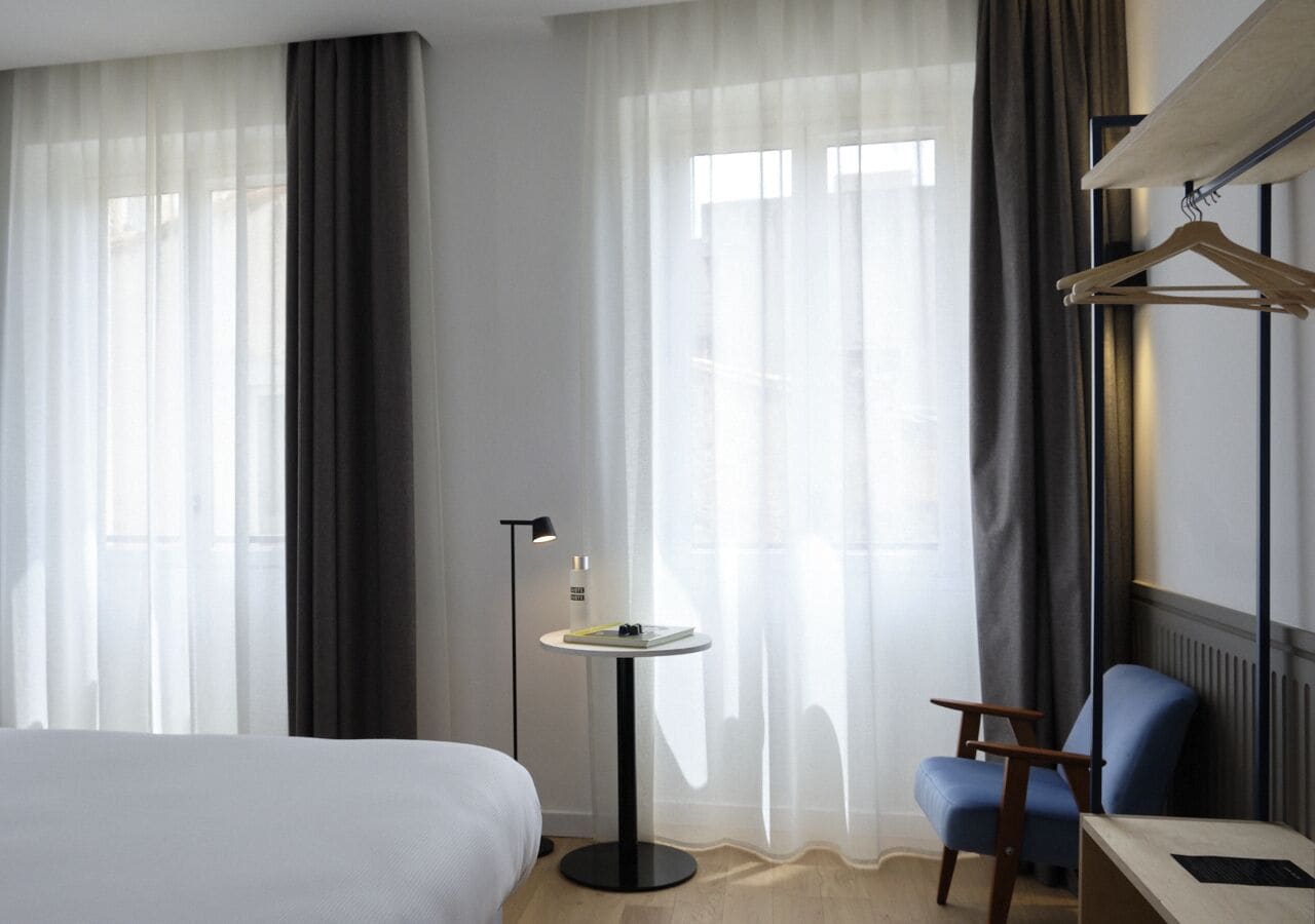 A hotel room with a bed, a small round table with a lamp, a chair, and a coat rack.