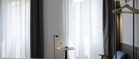 A hotel room with a bed, a small round table with a lamp, a chair, and a coat rack.