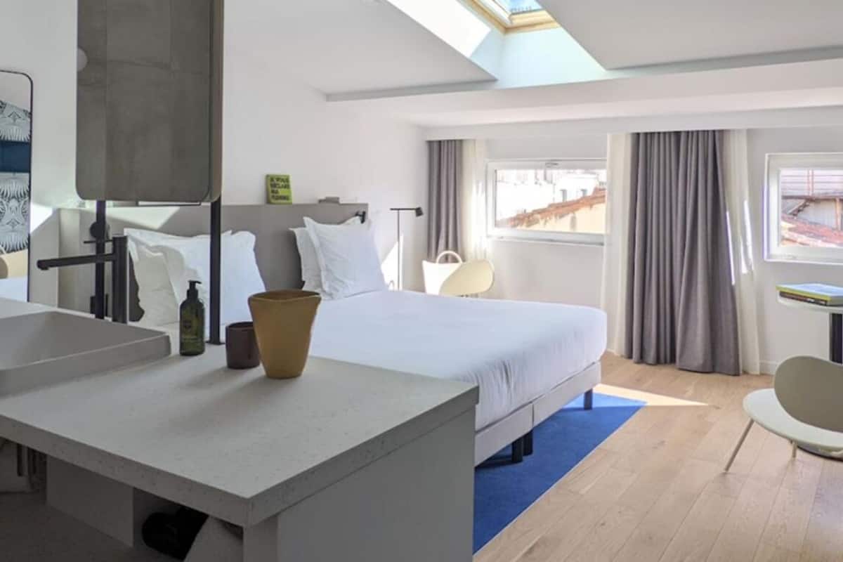 A modern hotel room with a large bed, a desk, a chair, a bathroom with a sink, and a TV mounted on the wall.