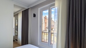 A hotel room with a bed, a window with curtains, a door, and a view of buildings outside. - Maison Juste (Marseille)