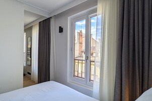 A hotel room with a bed, a window with curtains, a door, and a view of buildings outside.