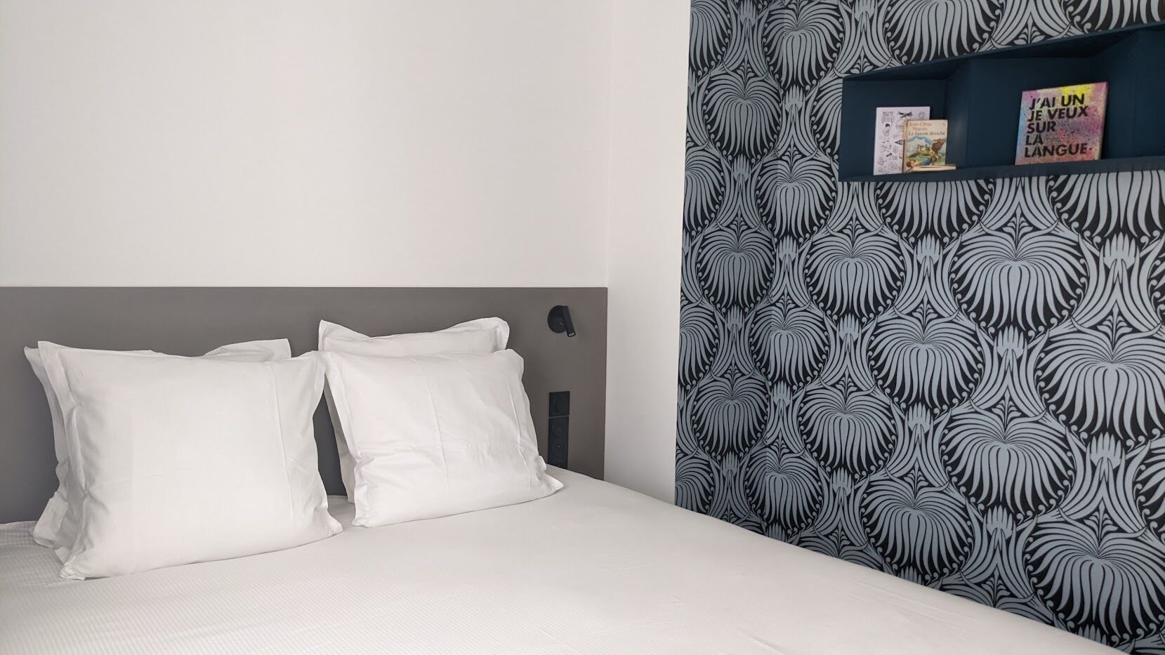 Un lit bien fait, avec du linge de lit blanc et des oreillers, une tête de lit sombre et un mur recouvert d’un papier peint à motifs noir et blanc.
