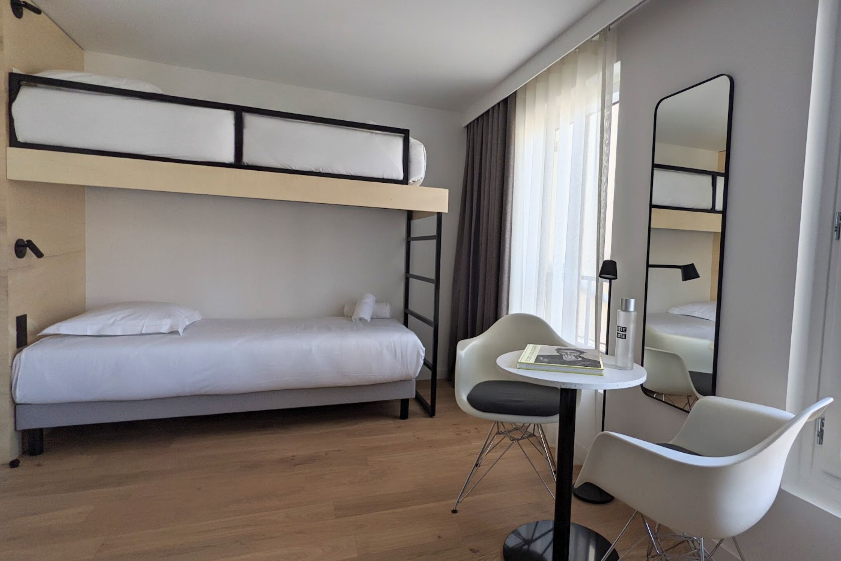 Ein Hotelzimmer mit Etagenbett, einem kleinen Tisch mit Stuhl, einem Spiegel und einem Fenster mit Vorhängen.