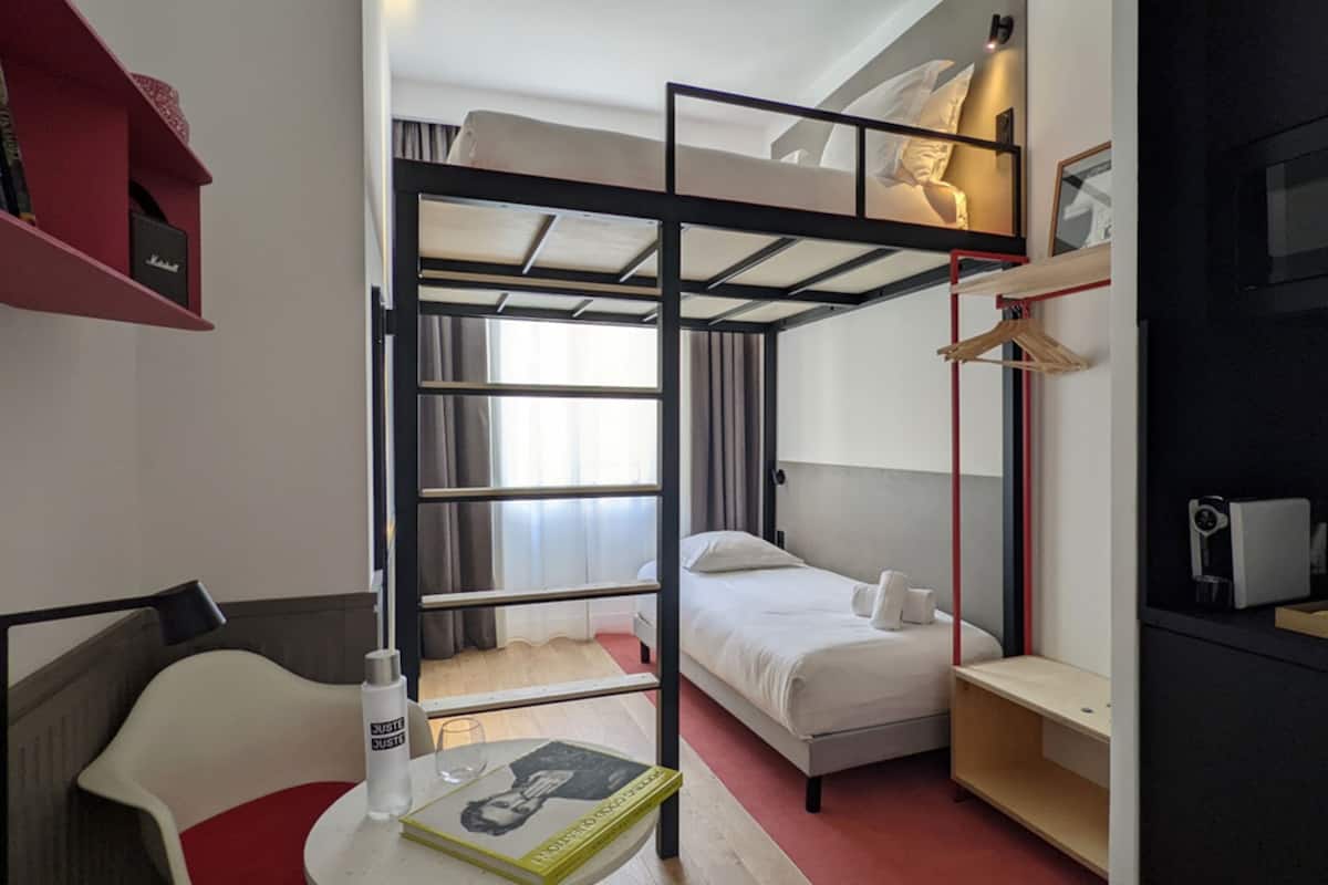 A compact hotel room with a bunk bed, a single bed, a desk with a chair, a small bookshelf, and a bedside table.