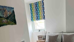Un dormitorio con cama, almohadas, una mesita de noche, un calentador, una ventana con cortinas y un cuadro en la pared.