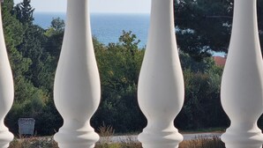 Ein Balkon mit weißen Brüstungen, der auf eine Küstenlandschaft mit Bäumen und dem Meer blickt.