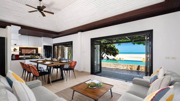 Ein modernes Wohnzimmer mit einer Couch, einem Couchtisch und einem Essbereich mit Stühlen und einem Tisch. Durch eine offene Tür ist Blick auf einen Pool und einen Strand.