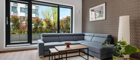 A modern living room with a grey sectional sofa, a wooden coffee table, and a large window offering a view of a building and trees.