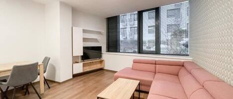 A modern living room with a wooden floor, a pink sofa, a wooden coffee table, a TV on a stand, and a window with blinds.
