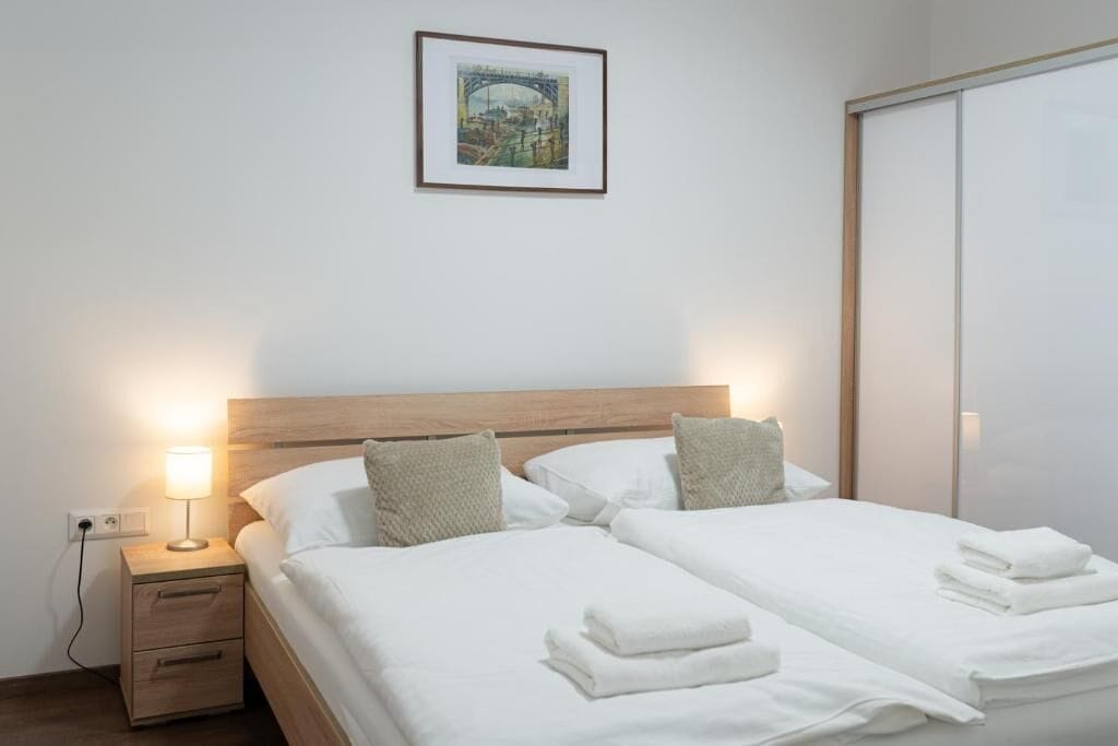 A hotel room with a wooden headboard, two beds with white linens, a nightstand with a lamp, a framed picture on the wall, and a sliding door closet.
