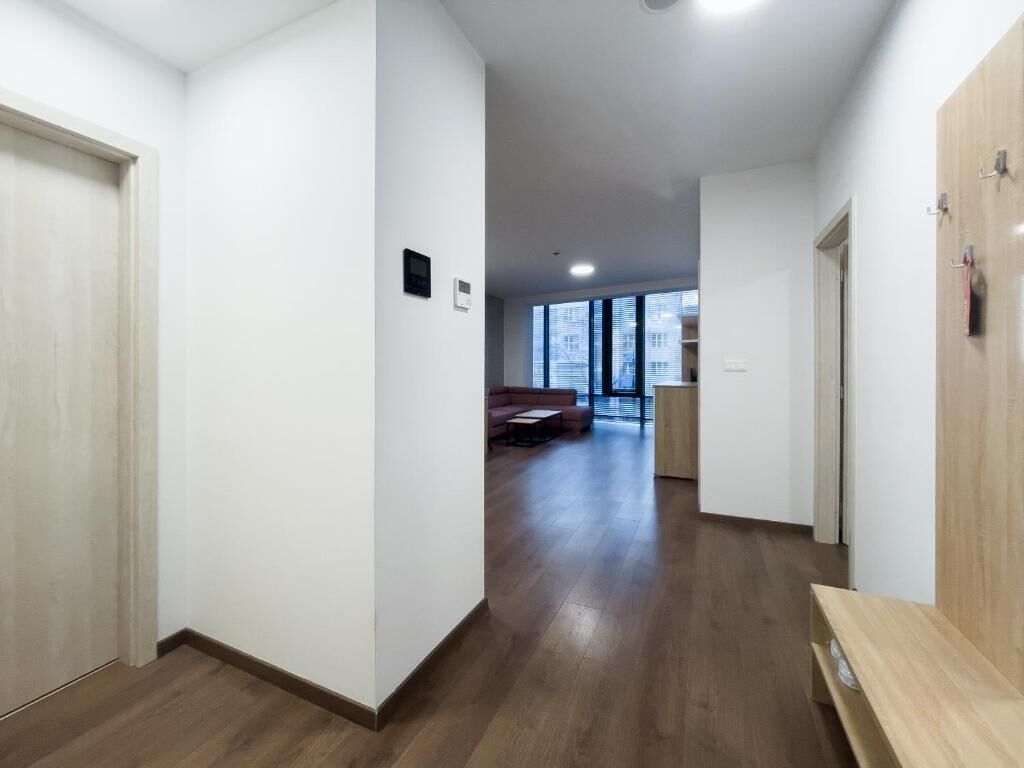 A hallway with wooden flooring, white walls, and a seating area with a sofa and table in the distance.