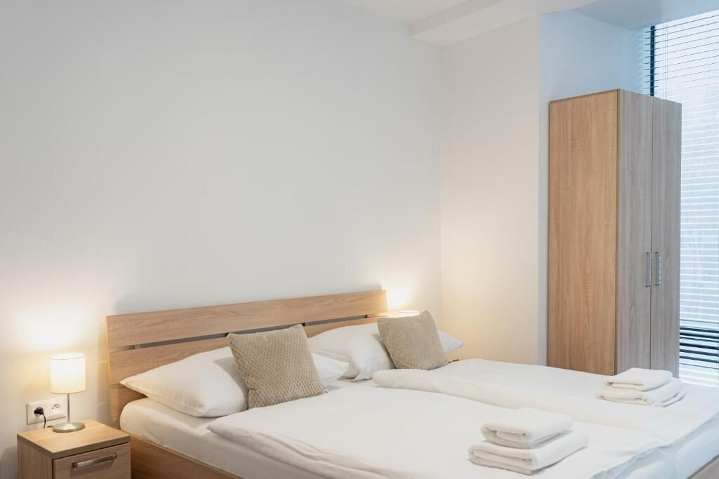 A modern hotel room with a wooden headboard bed, a nightstand with a lamp, and a wardrobe.