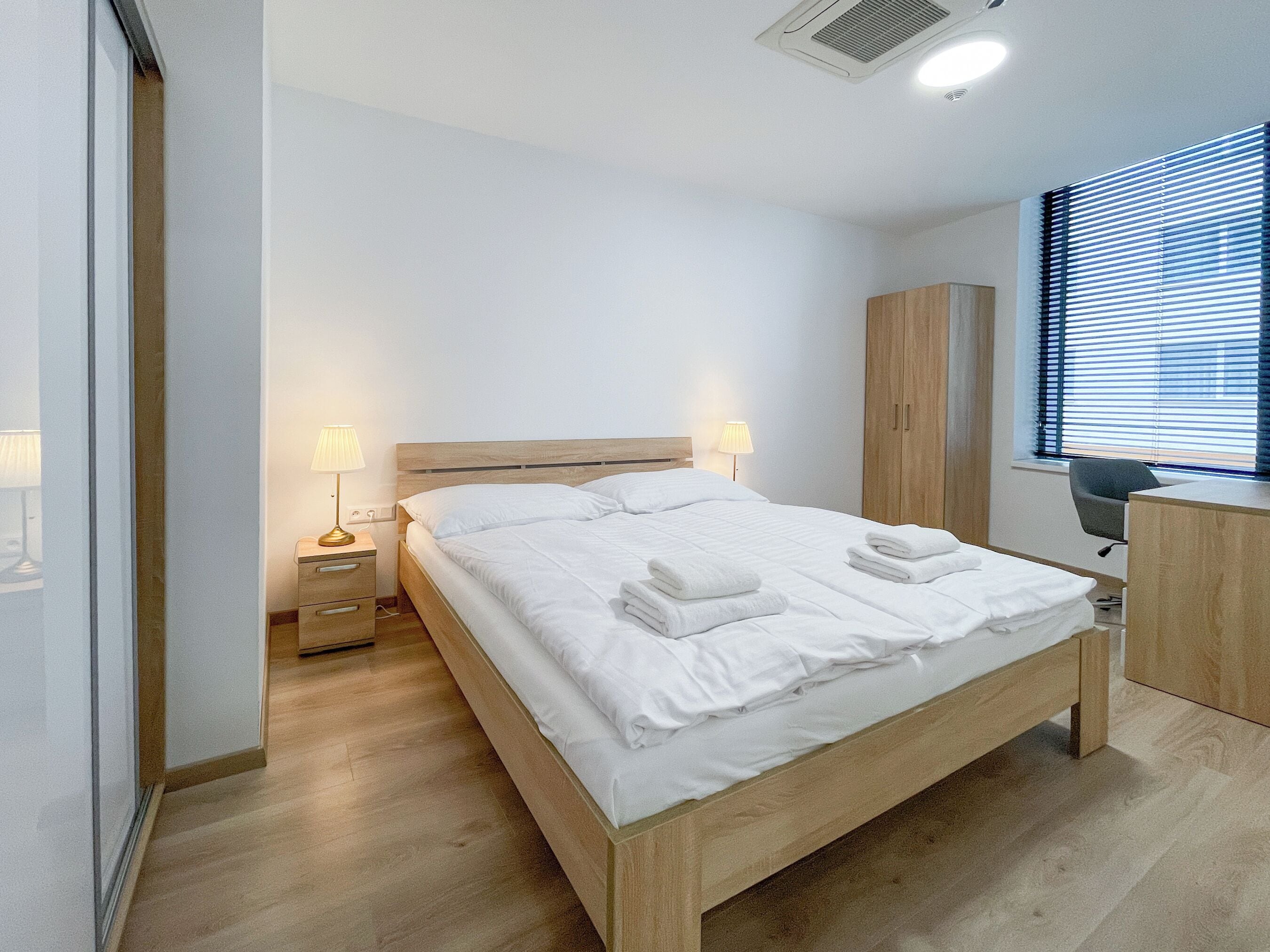 Una habitación de hotel moderna con una cama de madera, lámparas de noche, un escritorio y una ventana con persianas.