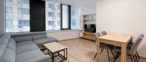 Una sala moderna con un sofá seccional gris, una mesa de centro de madera, una mesa de comedor con cuatro sillas y un televisor sobre un mueble de madera.