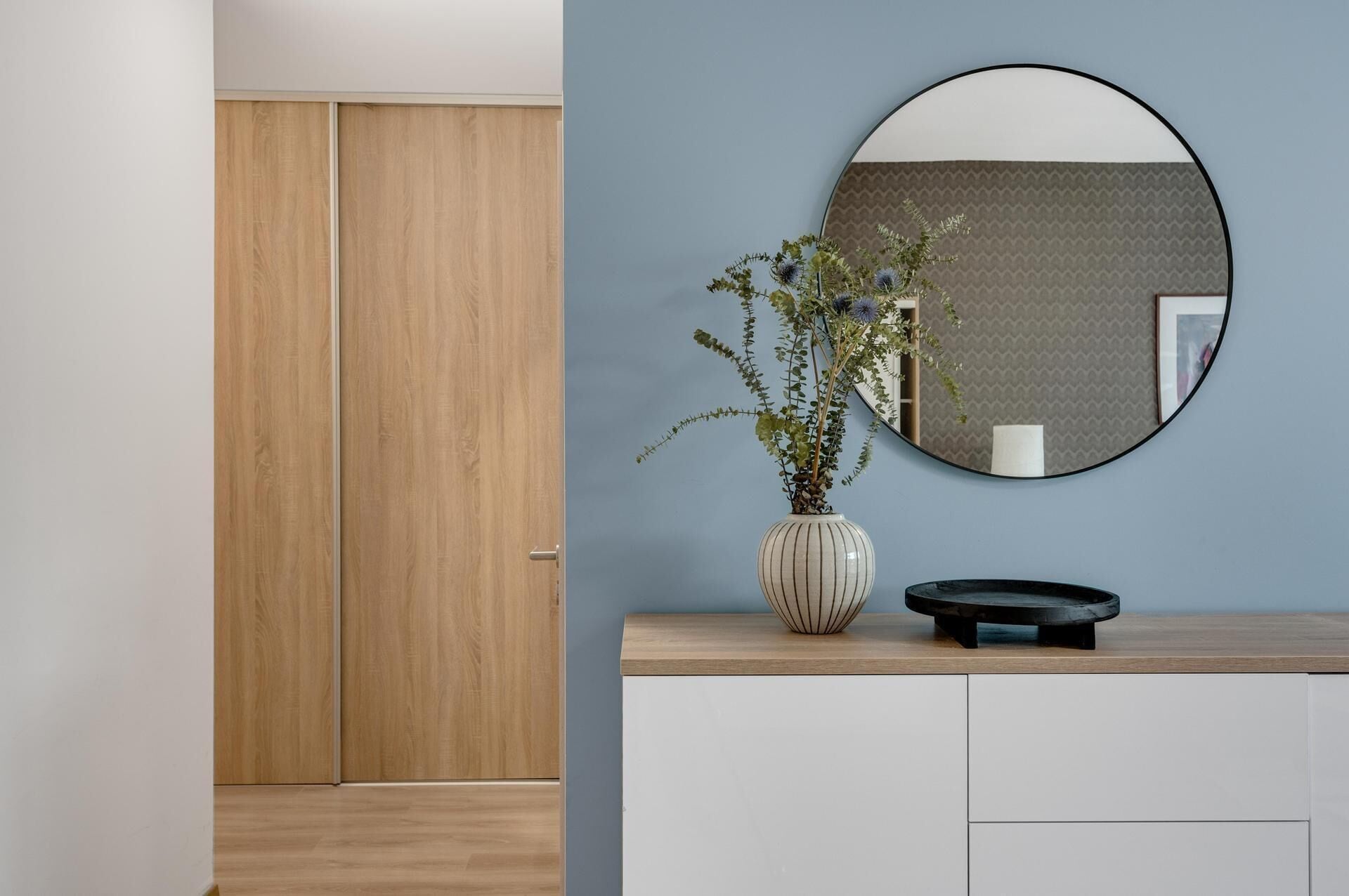 Une pièce avec une armoire en bois, une commode blanche ornée d’un vase de fleurs et un miroir rond accroché au mur.