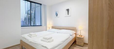 A modern bedroom with a wooden bed, a bedside table with a lamp, and a window with blinds.
