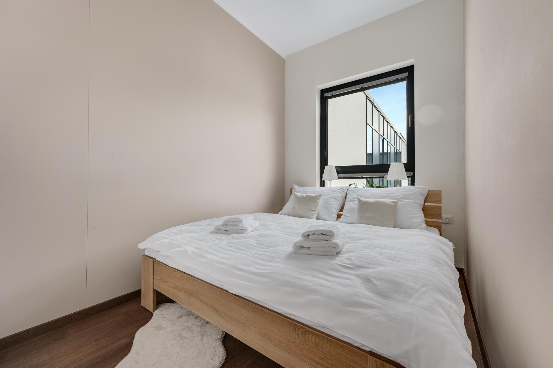 Un dormitorio moderno con una cama grande, ropa de cama blanca y vistas a un edificio a través de una ventana.