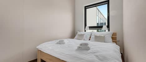 Un dormitorio moderno con una cama grande, ropa de cama blanca y vistas a un edificio a través de una ventana.