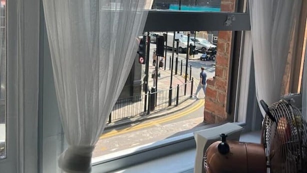 Una habitación con un ventanal grande, un ventilador y vistas a una calle con edificios y un autobús.