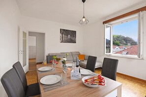Ein Essbereich mit einem Tisch für vier Personen, einer Couch im Hintergrund und einem großen Fenster mit Blick auf einen Hügel und Gebäude.