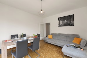 Un coin repas avec une table dressée pour deux personnes, un canapé gris agrémenté de coussins orange et une œuvre d’art en noir et blanc accrochée au mur.