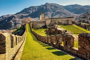 A historic stone wall with a grassy pathway leading to a fortified structure with towers, set against a mountainous backdrop.