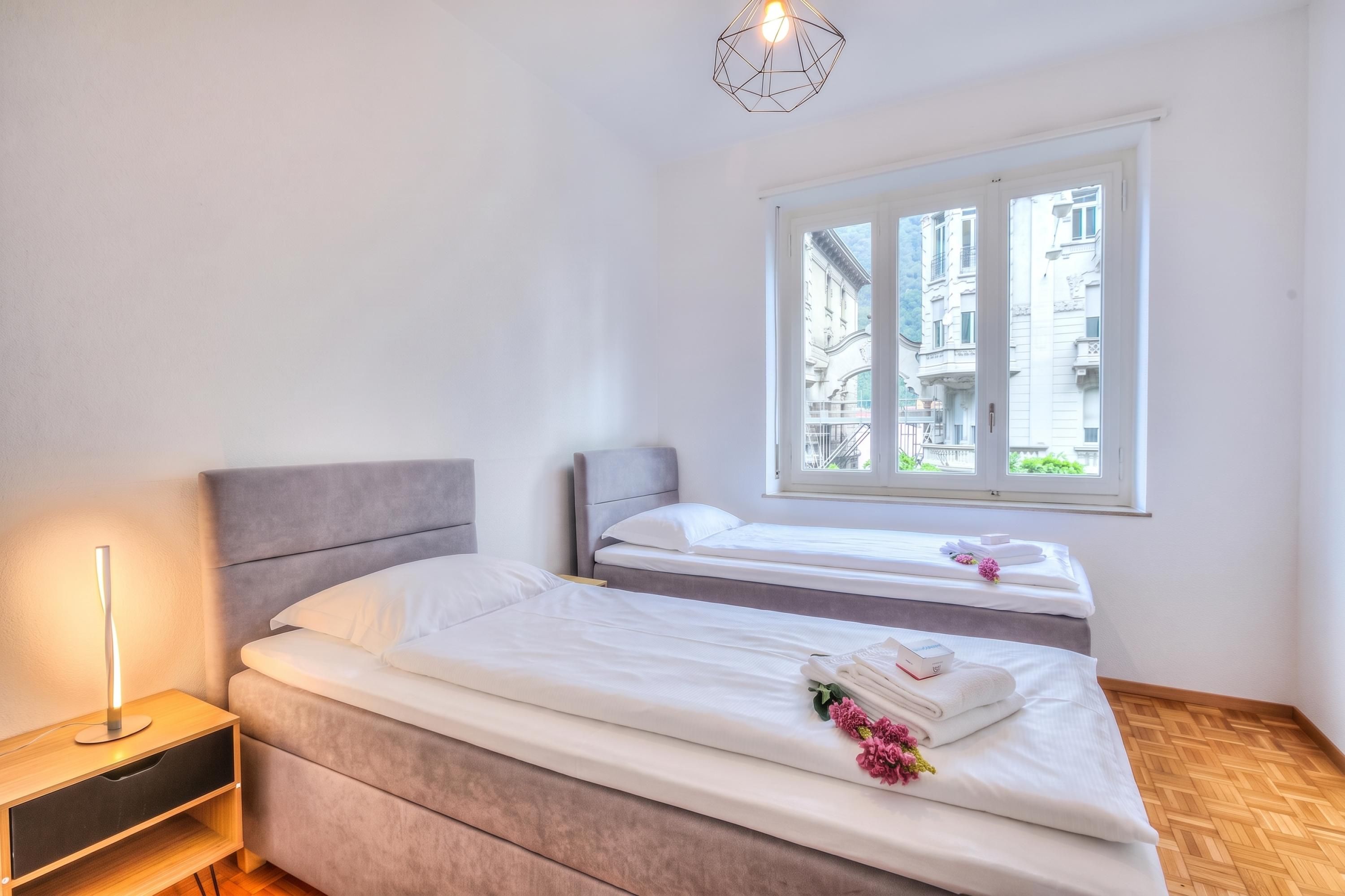 A hotel room with two beds, a bedside table, a lamp, and a window with a view of buildings.