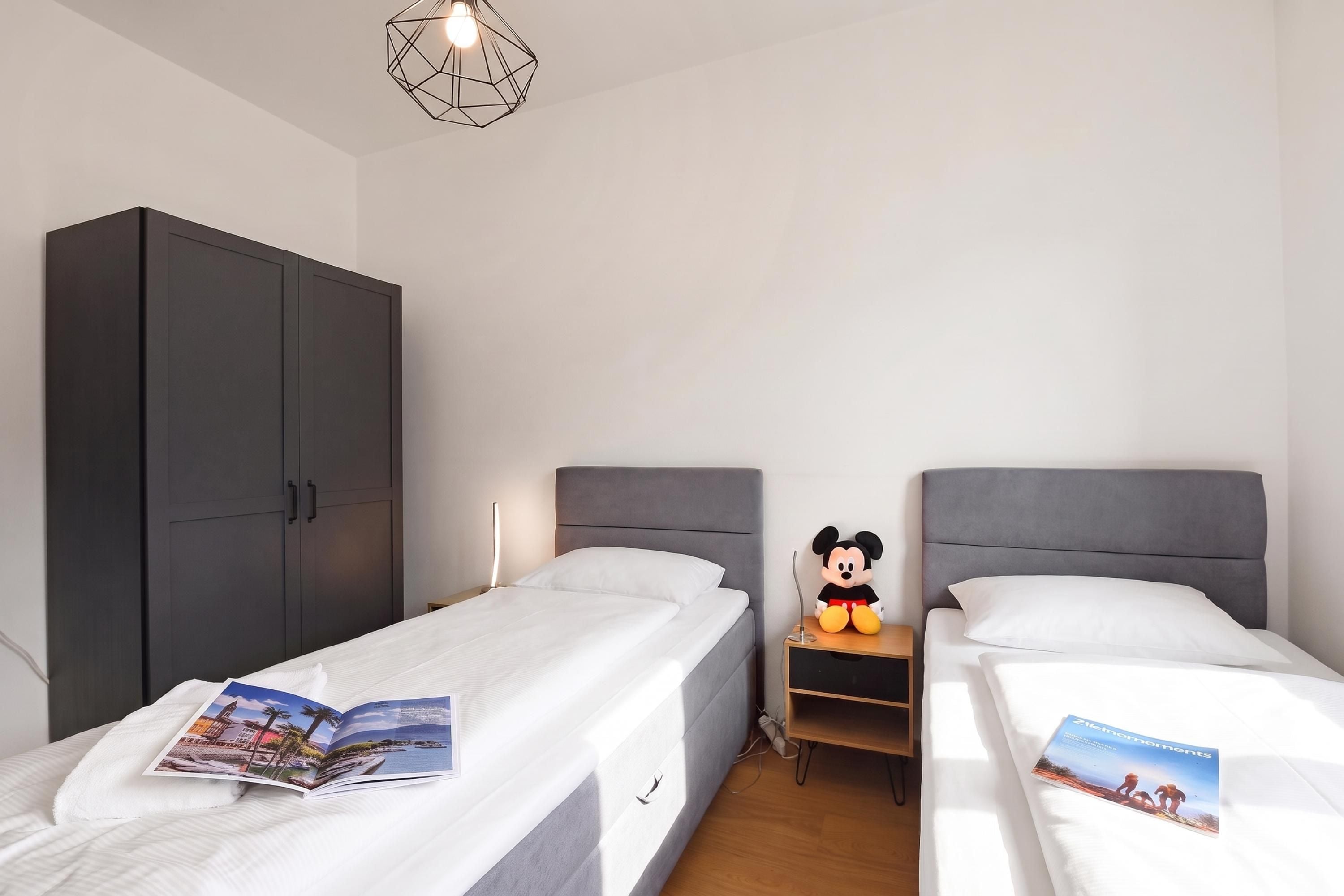 A hotel room with two single beds, a wardrobe, a Mickey Mouse plush toy, and a magazine on the beds.