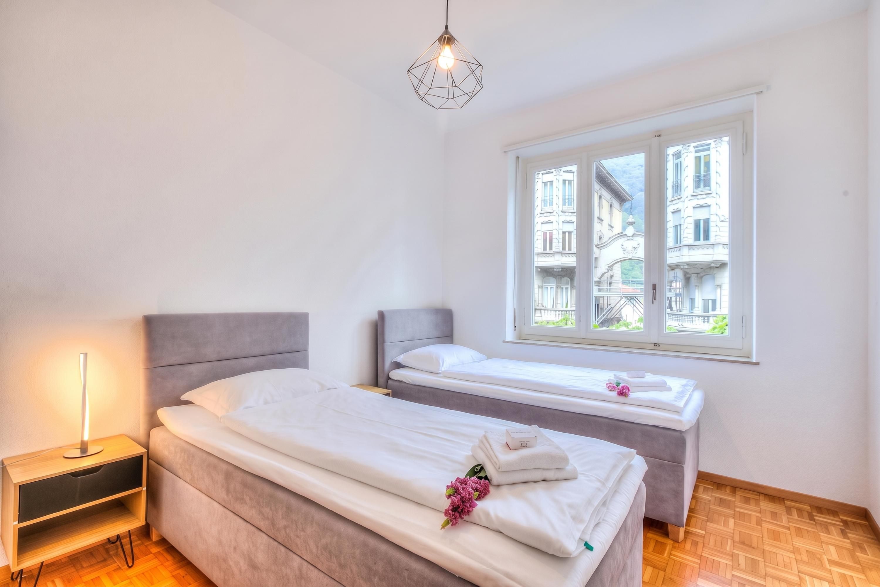Een hotelkamer met twee bedden, een nachtkastje, een lamp en een raam met uitzicht op gebouwen.