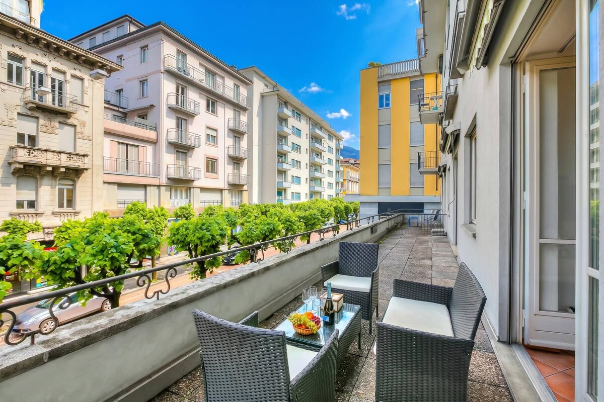 A balcony with wicker furniture, a table with a bottle and fruit, and a row of buildings with balconies.