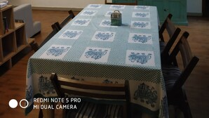 Un coin repas avec une table recouverte d’une nappe à motifs, des chaises, un ventilateur de plafond, une étagère murale avec des bouteilles et un téléviseur sur le côté gauche.