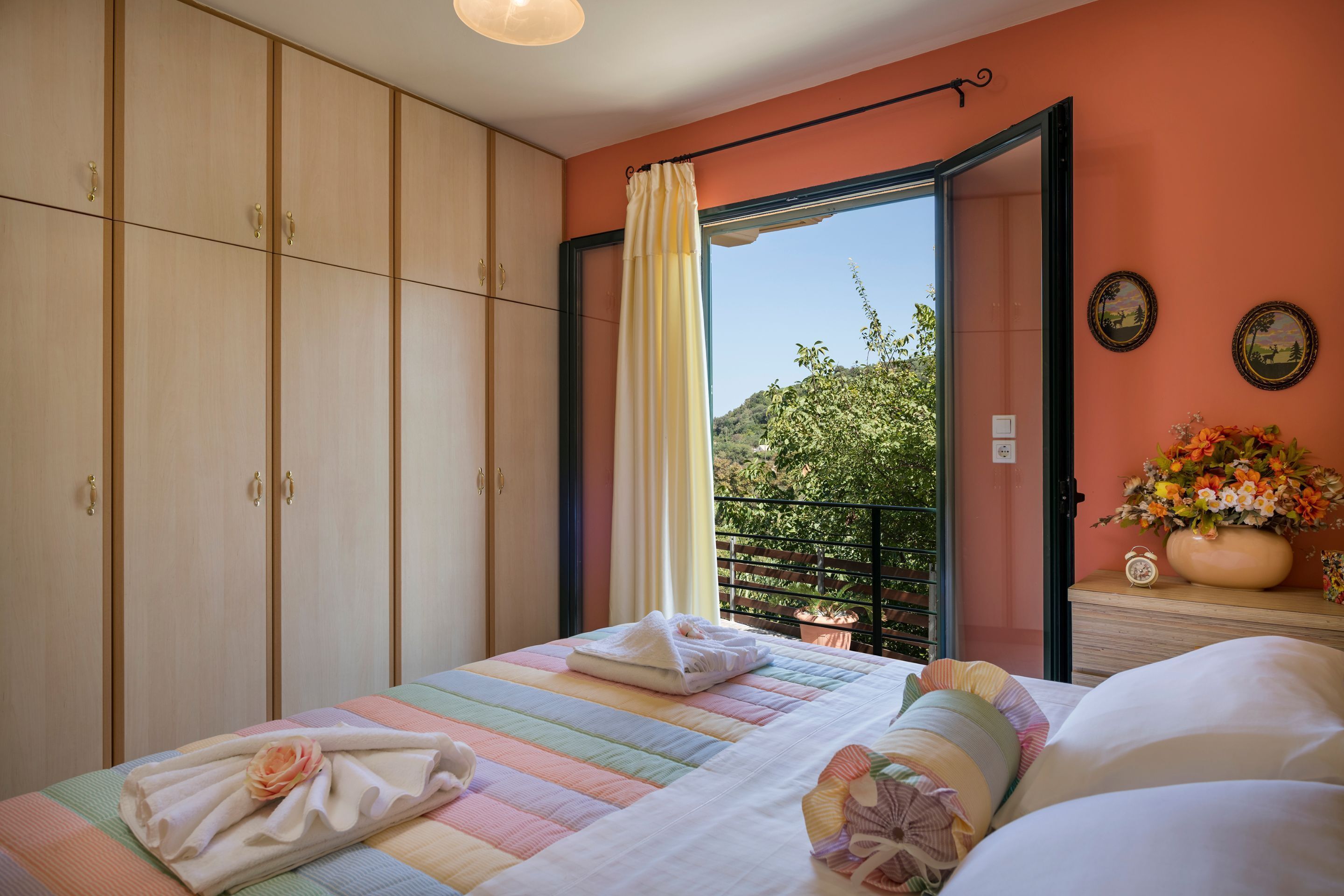 A bedroom with a bed, a wardrobe, a window with curtains, a nightstand with a clock and flowers, and a view of greenery outside.